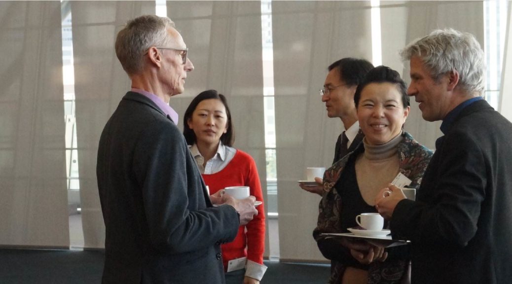 Five people are to be seen. One of them is a speaker of the symposium. He is holding a businesscard. The other people are holding cups of coffee and one woman next to the speaker is smyling towards the viewer